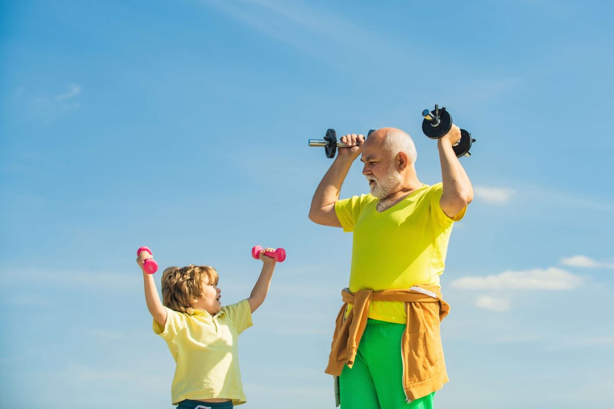 Grandpa lifting dumbbells for exercise with his grandson in front of blue skies.