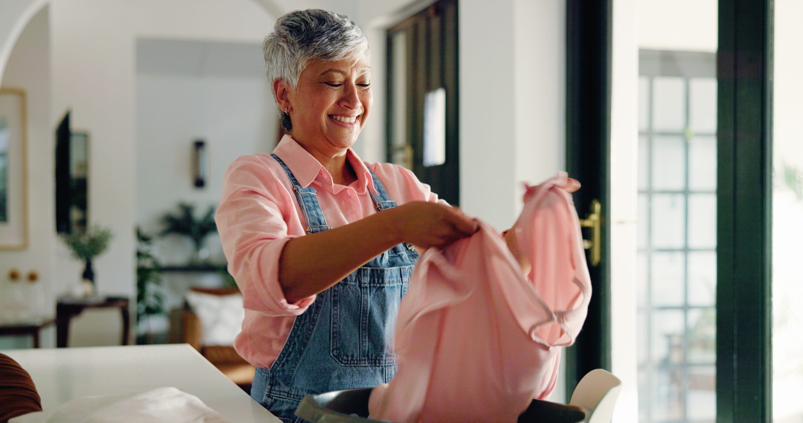 Happy old woman, cleaning and folding laundry in home for housekeeping or hygiene maintenance. Housework, person and fabric in basket for chores, morning routine and organizing clothes in retirement